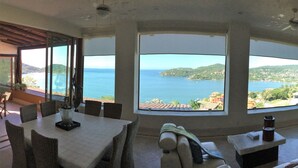 A room with a large panoramic window offering a view of the sea and hills. There is a dining area with chairs and a table, and a seating area with a sofa and a small table. - Spectacular luxury penthouse at La Ropa beach, unique view, private rooftop pool (Playa la Ropa)