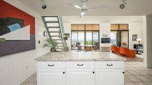 A modern kitchen with a granite countertop, white cabinets, and a built-in stove. There is a dining area with a table and chairs, and a living room with a sofa and a TV. A ladder is propped against the wall, and a painting is on the wall. - Exclusive Key West Beach Front - 2BR Luxury Condo w/Gym and Pool Access (Newly Refurnished!) (Key West)