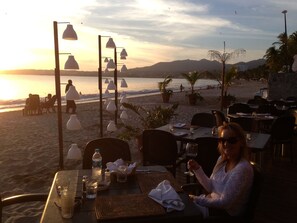 Ein Restaurant direkt am Strand mit Außenbereich, Tischen und Stühlen mit Blick auf das Meer und die Berge.