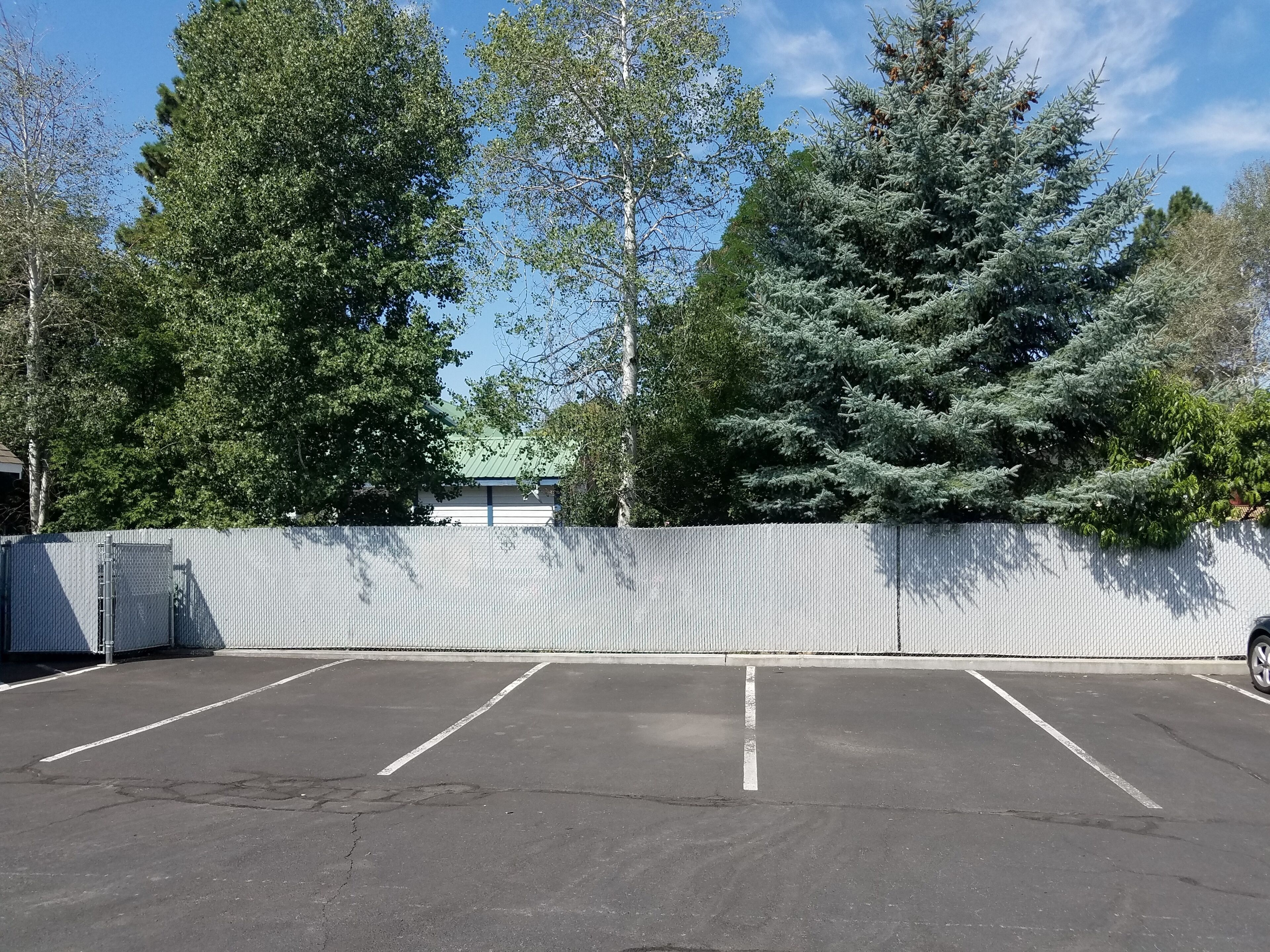 A parking lot with a fenced area, trees, and a building with a green roof in the background.