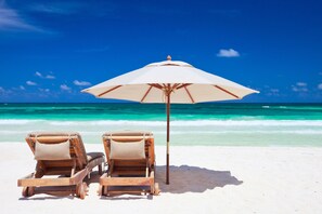 Two wooden lounge chairs with beige cushions and a large white umbrella on a sandy beach with clear turquoise water and a blue sky. - SNOWBIRD SPECIAL / Golf Course Villa-2BR/ Lake View / PET OK/Short Walk to Beach (Miramar Beach)