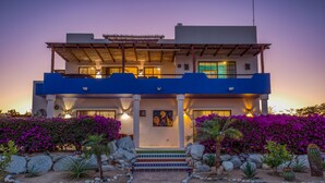A two-story building with a balcony, a well-lit entrance, and a garden with rocks and plants. - Sweeping ocean views steps to beach w/pool & Firepit. Close to Tres Palapas!  (Los Barriles)