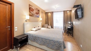 Habitación de hotel con una cama grande, un escritorio con una silla, un televisor fijado a la pared y una ventana con cortinas.