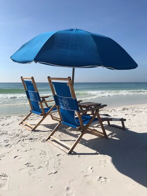 Zwei Liegestühle unter einem blauen Sonnenschirm an einem sandigen Strand mit Fußabdrücken.