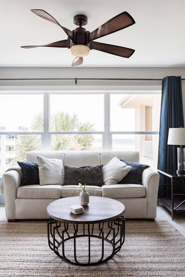 A living room with a ceiling fan, a white sofa with cushions, a round coffee table, and a view of the outdoors through large windows.