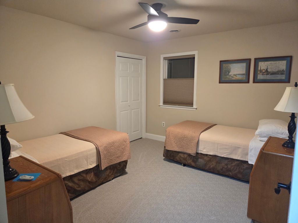 A bedroom with two beds, a ceiling fan, a window with blinds, and a door.