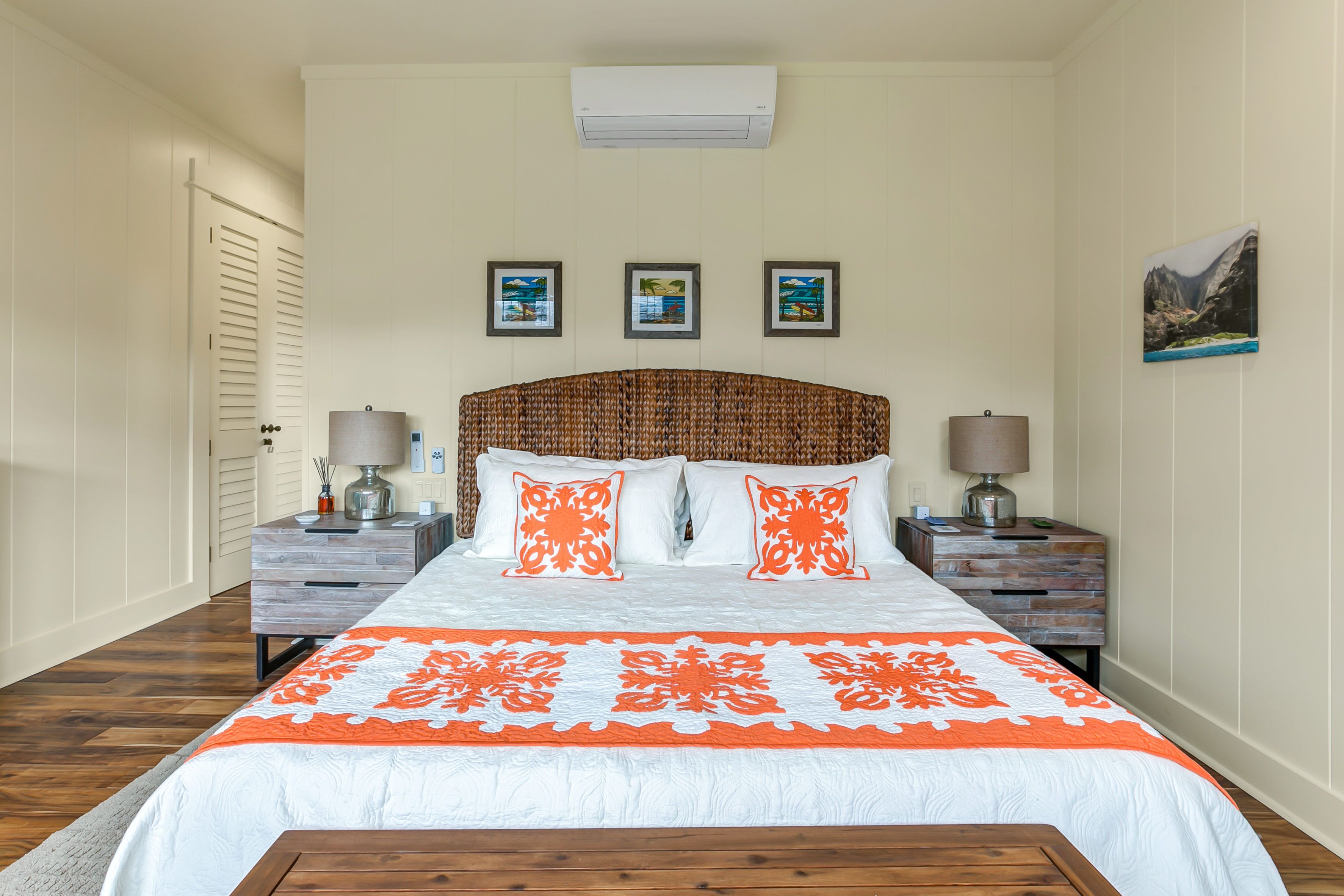 Une chambre avec un grand lit, deux tables de chevet et un banc en bois. Trois tableaux encadrés ornent le mur, et un appareil de climatisation est installé au-dessus du lit.