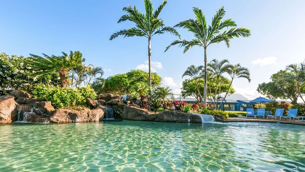 Una piscina cristalina con cascada, rodeada de palmeras y un edificio con sillas azules.