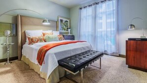 A bedroom with a bed, bedside lamps, a bench, a nightstand, a dresser, and a window with curtains. - Oakwood at Mosaic South End (Charlotte)