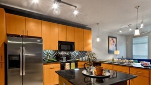 A modern kitchen with wooden cabinets, a stainless steel refrigerator, a microwave, and a black countertop island. - Oakwood at Mosaic South End (Charlotte)