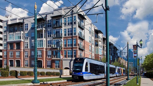A modern train station with a high-speed train, multi-story buildings, and overhead electrical lines. - Oakwood at Mosaic South End (Charlotte)