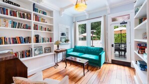 A cozy living room with a teal sofa, wooden flooring, and white walls. There are bookshelves filled with books, a small table with a lamp, and a door leading to an outdoor area with umbrellas.