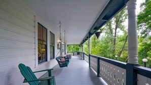 A covered porch with a swing chair and a dining table, surrounded by greenery.
