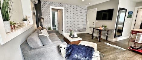 A modern living room with a grey sectional sofa, a blue furry ottoman, and a dining area with a glass table and chairs.
