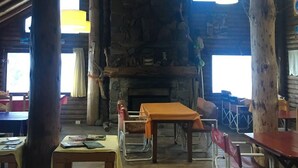Ein rustikaler Innenraum mit Kamin aus Stein, Holzbalken und hoher Decke. Tische und Stühle sind zum Essen angeordnet.