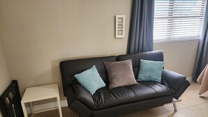Un canapé en cuir noir avec trois coussins, une petite table d’appoint blanche et un tableau encadré au mur.