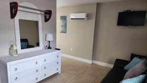 Un salon avec une commode blanche, un canapé noir, un téléviseur à écran plat et un climatiseur fixé au mur.