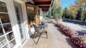 Une terrasse avec une table et des chaises, un parterre de fleurs et une vue sur des arbres et une route.