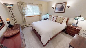 Une chambre à coucher comprenant un lit, des tables de chevet, une commode, une lampe, une fenêtre avec des rideaux, un tableau encadré au mur et un miroir.