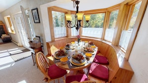 A round wooden table set for dining with chairs, a chandelier, and a view of trees through large windows.