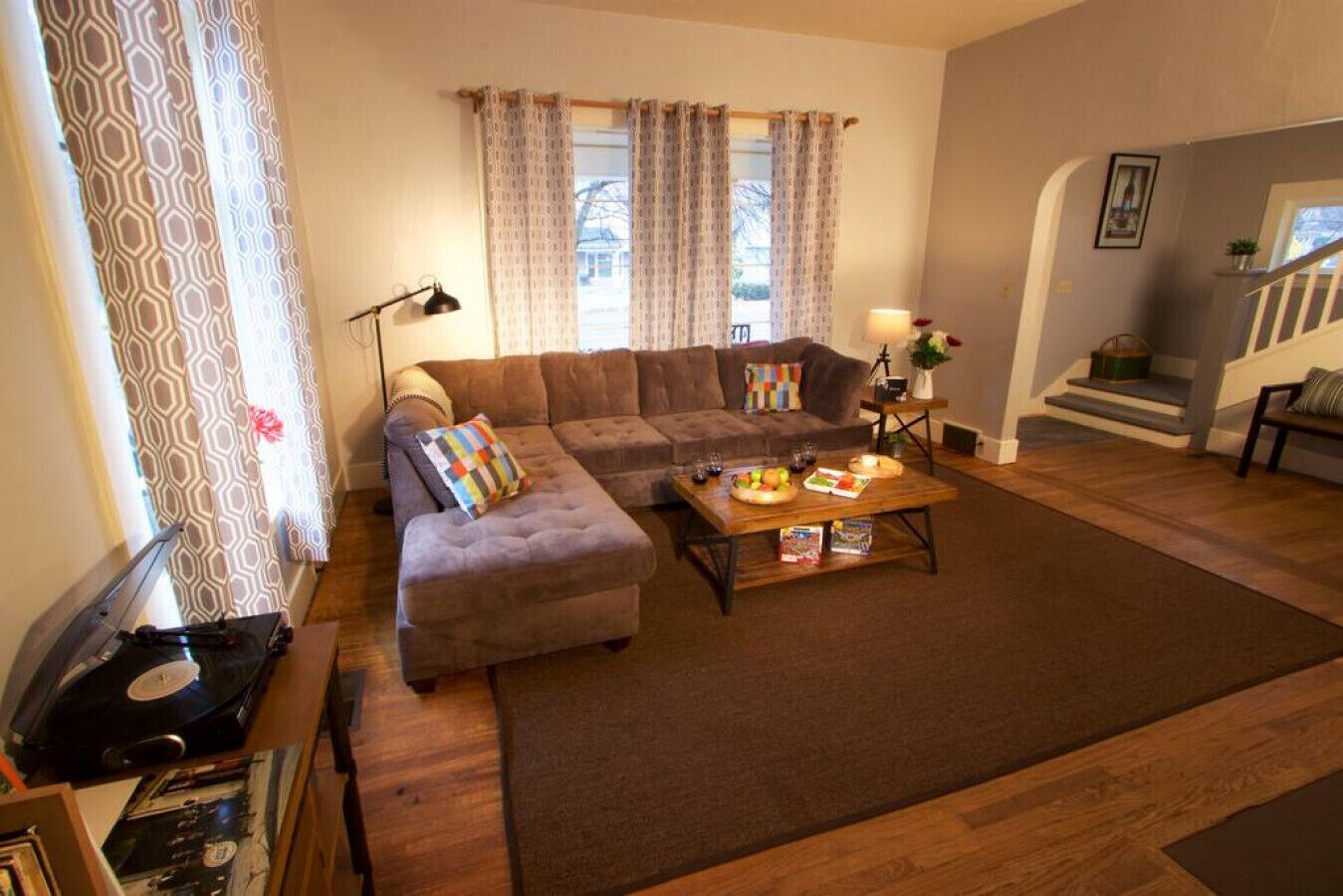 A living room with a sectional sofa, a coffee table with snacks, a record player, and patterned curtains.