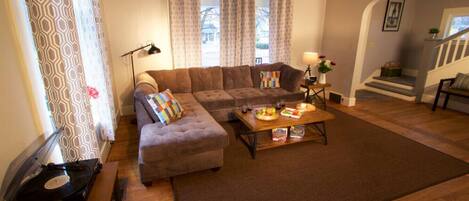 A living room with a sectional sofa, a coffee table with snacks, a record player, and patterned curtains.