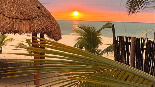 A beach scene with a thatched-roof hut, palm trees, and a sunset over the ocean. - Posada Maria Isabel (Celestún)