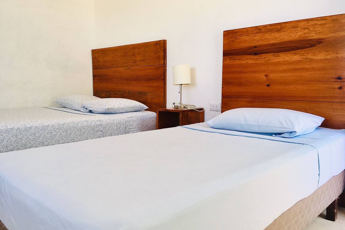 A hotel room with two single beds, each with a wooden headboard, a bedside table with a lamp, and a power outlet.