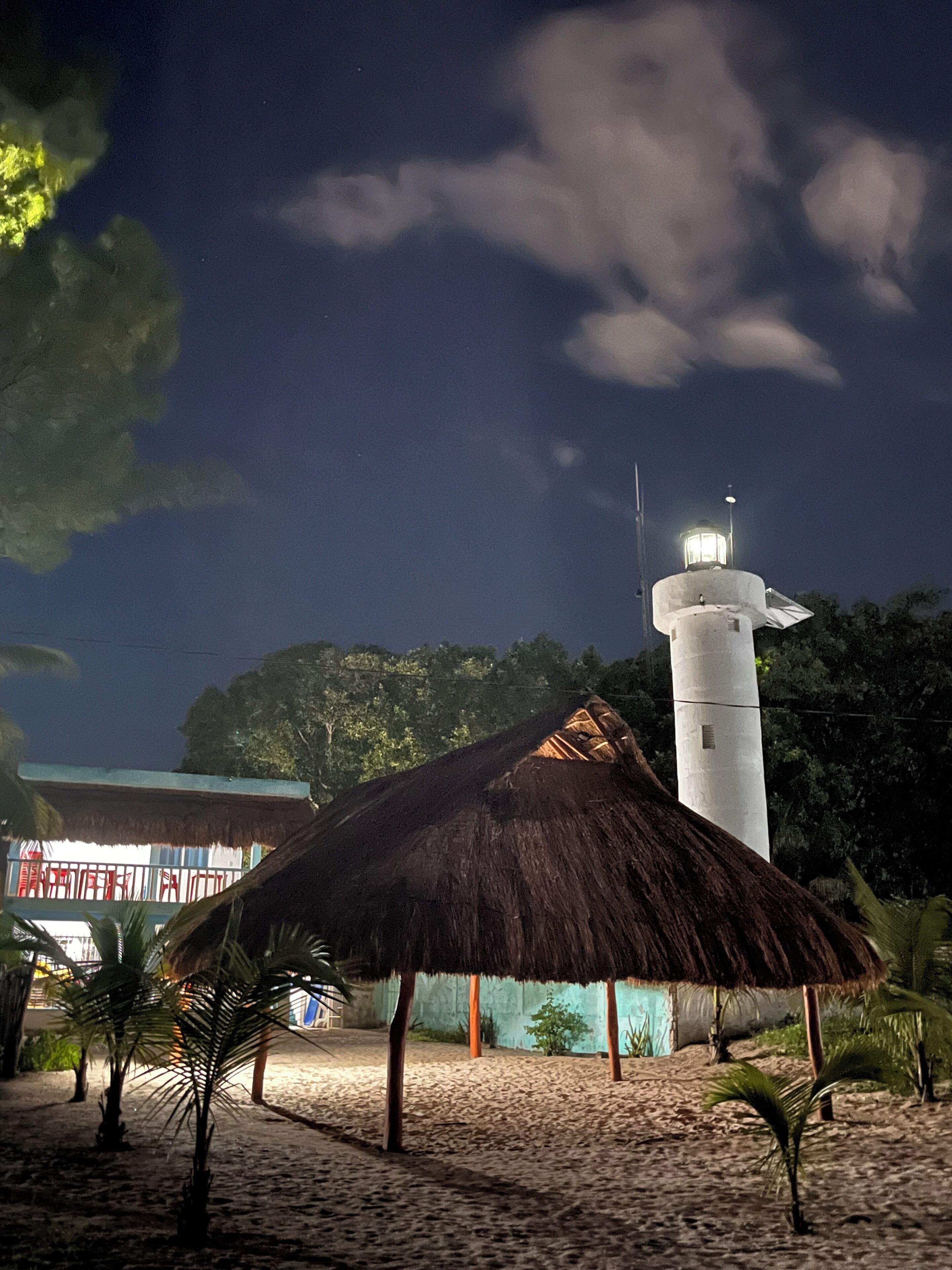 A lighthouse with a thatched roof structure nearby, surrounded by palm trees and a building with a blue exterior.