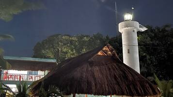 Um farol com uma estrutura de telhado de palha por perto, cercado por palmeiras e um edifício com fachada azul.