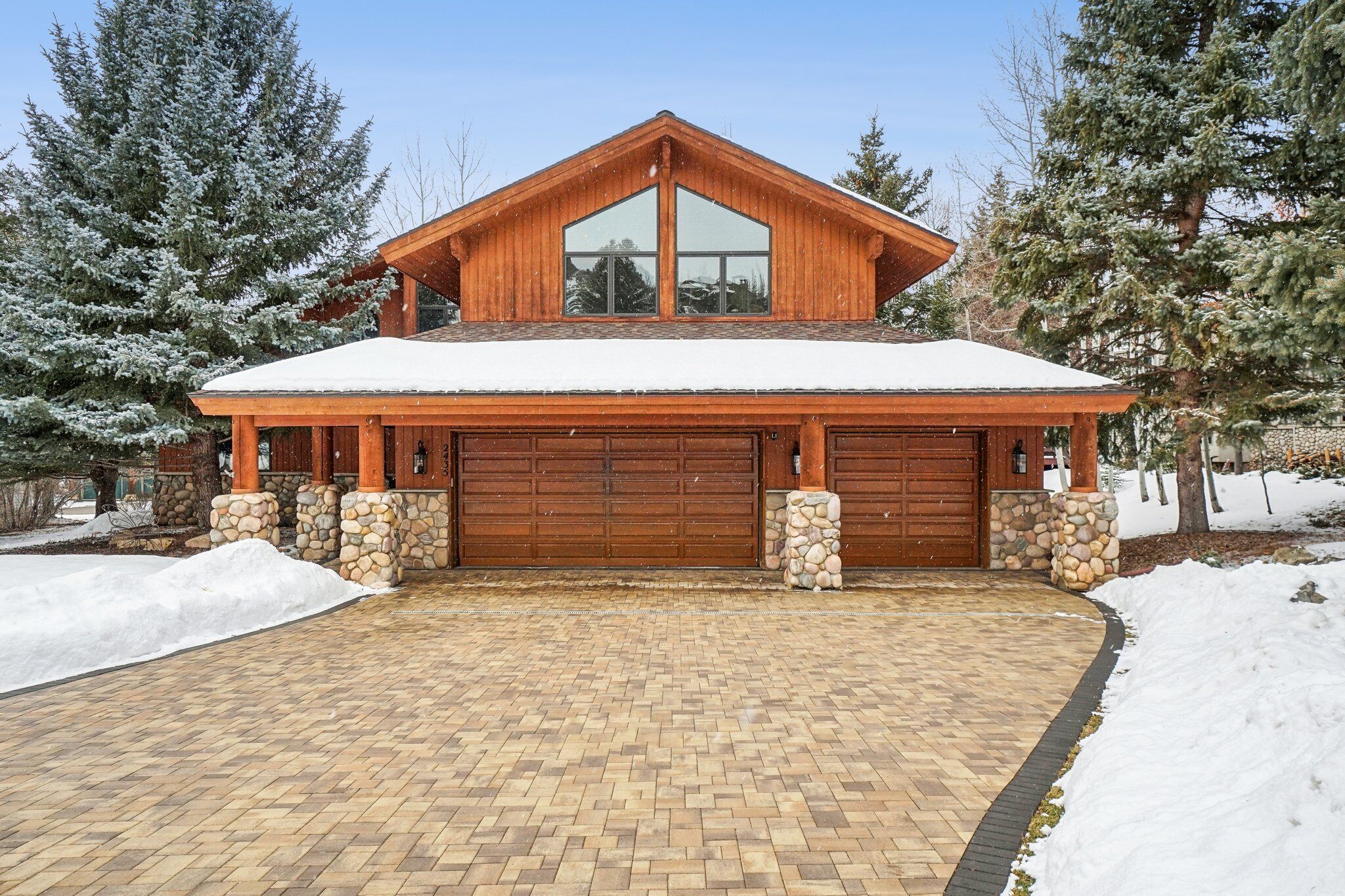 A two-story wooden house with a large garage, stone pillars, and a snow-covered roof.