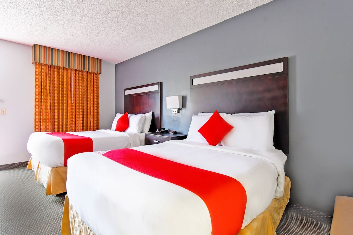 A hotel room with two beds, each with white bedding and red accents, a wooden headboard, a nightstand, and a mirror.