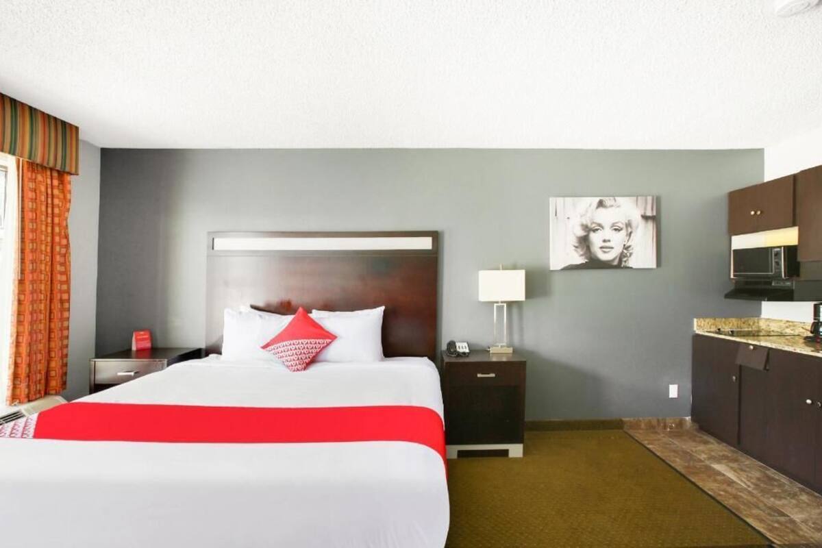 A hotel room with a bed, a nightstand, a lamp, a painting on the wall, and a kitchenette.