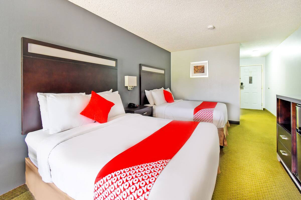 A hotel room with two beds, each with white bedding and red accents, a bedside table with a lamp, and a large mirror above the bed.