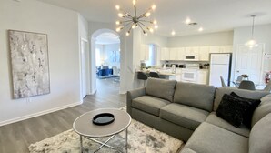 A modern living room with a grey sofa, a small round table, and a fluffy rug. There is a kitchen in the background with white cabinets and a refrigerator. - Family Paradise w/ Pvt Pool, BBQ Grill, Themed Bedrooms, Game Room & Club access (Kissimmee)