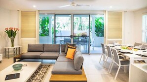 A modern living room with a grey sofa, a dining table with chairs, and a view of a garden through large glass doors. - Villa Del Mar - Beachfront Port Douglas (Port Douglas)