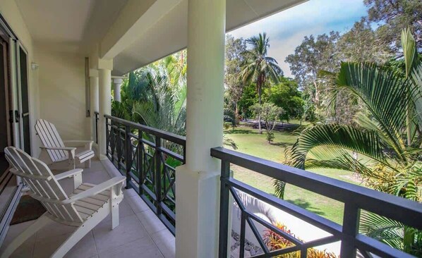 A balcony with two Adirondack chairs, a railing, and a view of a well-maintained garden with palm trees and a lawn.