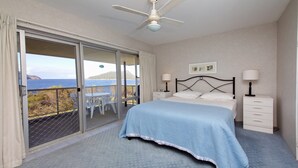 A bedroom with a bed, two bedside tables, a ceiling fan, and a view of the sea through a sliding glass door. - Shoalhaven 6 with Stunning Ocean Views (Nelson Bay)