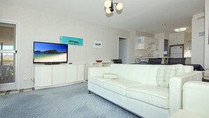 A living room with a white sofa, a flat-screen TV on a stand, and a kitchen in the background. - Shoalhaven 6 with Stunning Ocean Views (Nelson Bay)