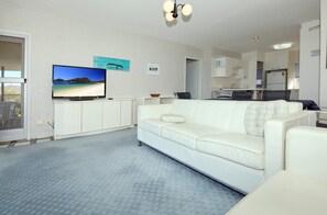 A living room with a white sofa, a flat-screen TV on a stand, and a kitchen in the background. - Shoalhaven 6 with Stunning Ocean Views (Nelson Bay)