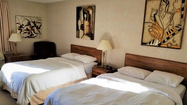 A hotel room with two beds, a wooden headboard, a nightstand with a lamp, and abstract wall art. - Abby Inn (Abbotsford)