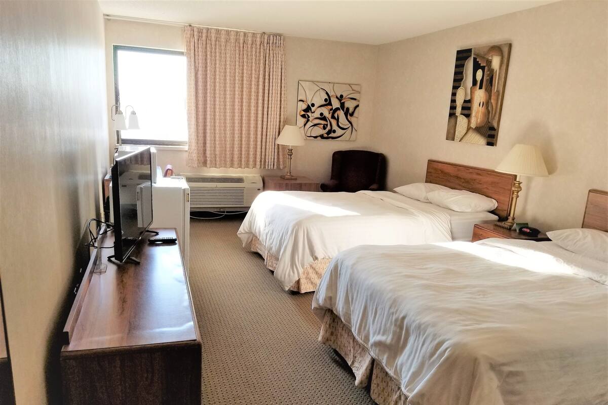 A hotel room with two beds, a desk with a computer, a TV, a window with curtains, an air conditioner, and artwork on the walls.