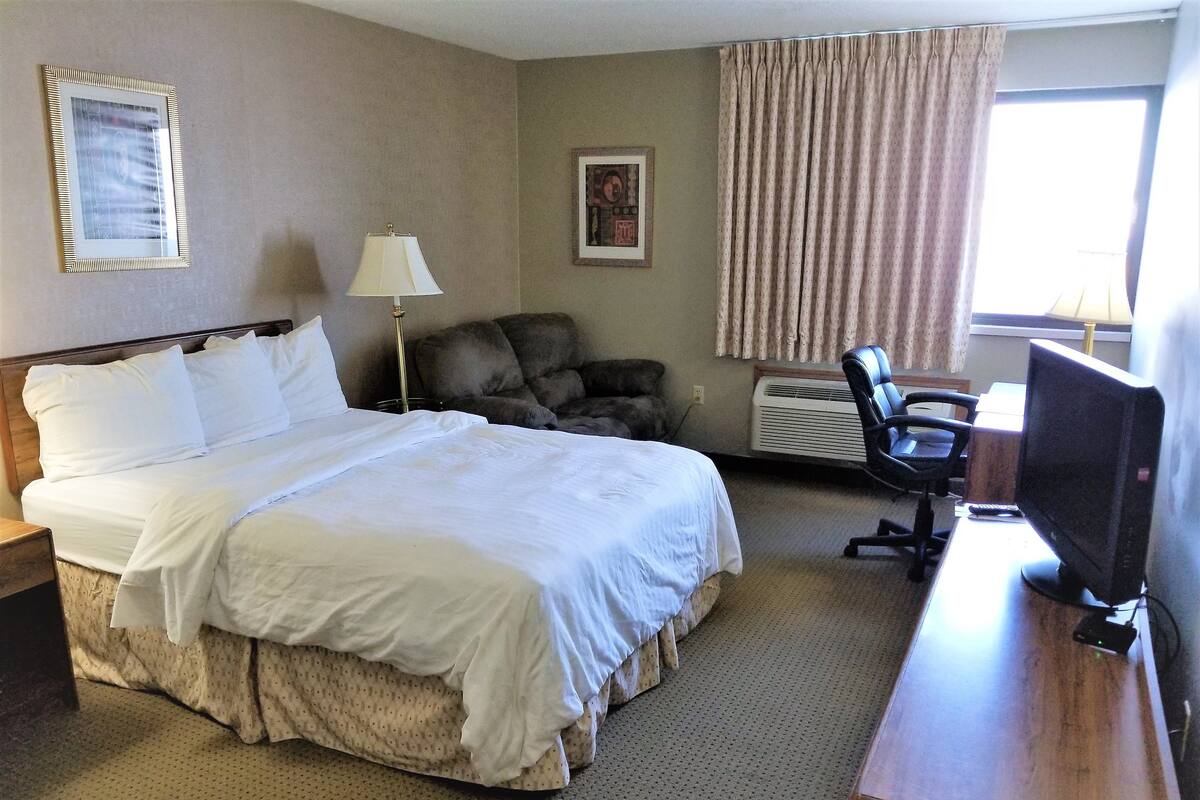 A hotel room with a bed, a desk with a computer, a chair, a lamp, and a window with curtains.