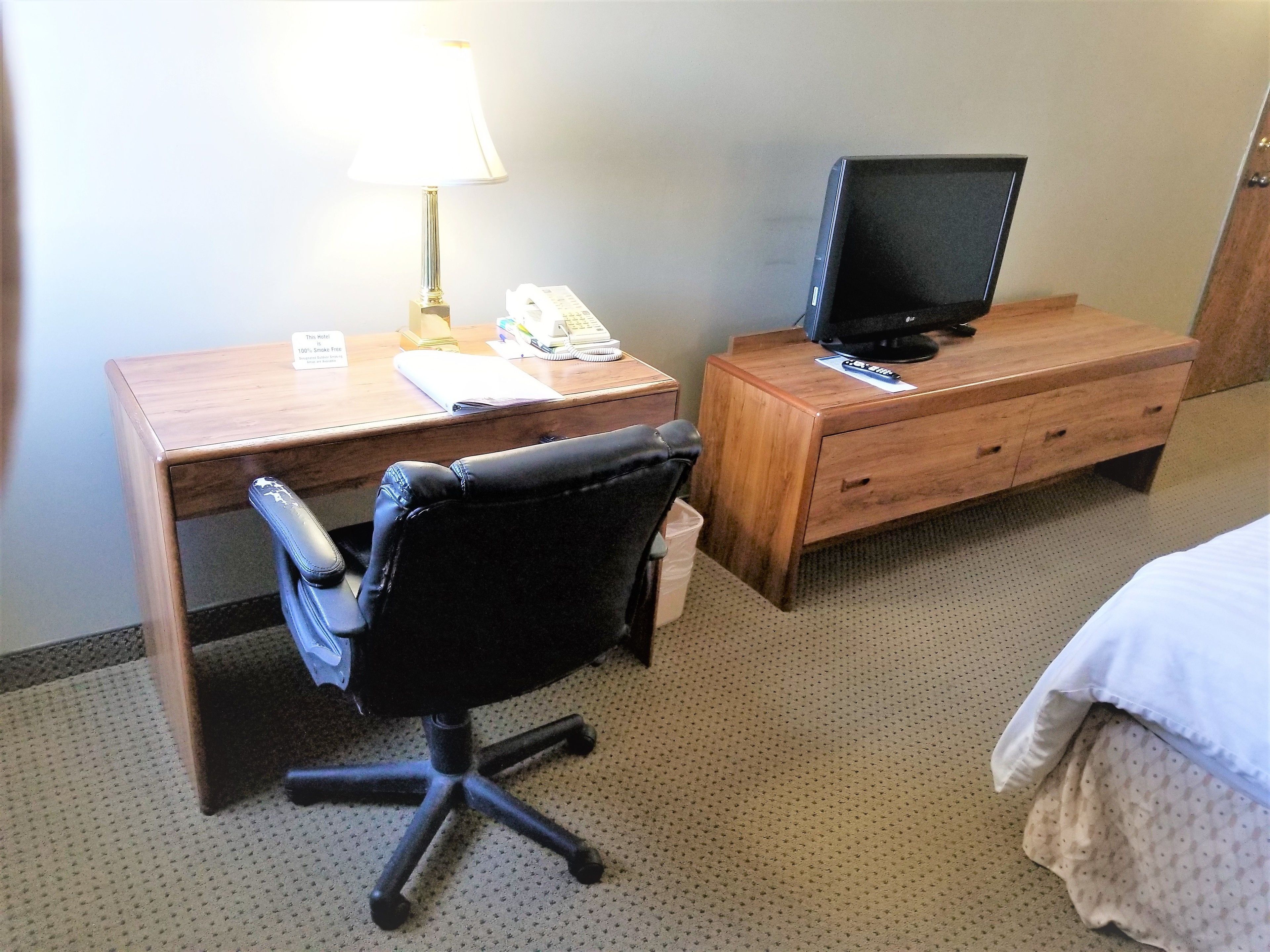 Een hotelkamer met een houten bureau, een zwarte bureaustoel, een televisie op een houten dressoir en een bed met wit beddengoed.