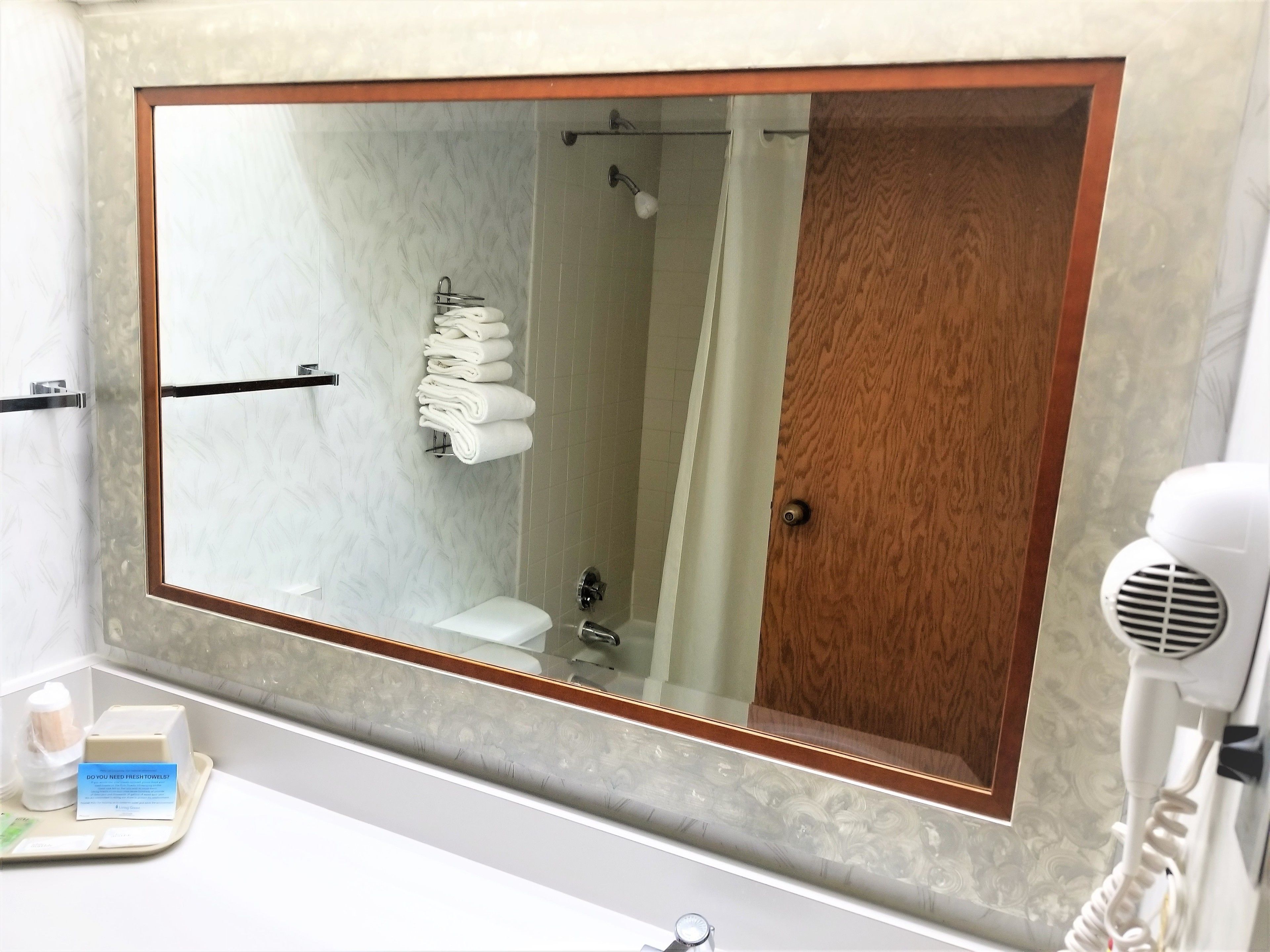 Une salle de bain avec un grand miroir, un porte-serviettes avec des serviettes pliées, un espace douche et un sèche-cheveux posé sur le comptoir.