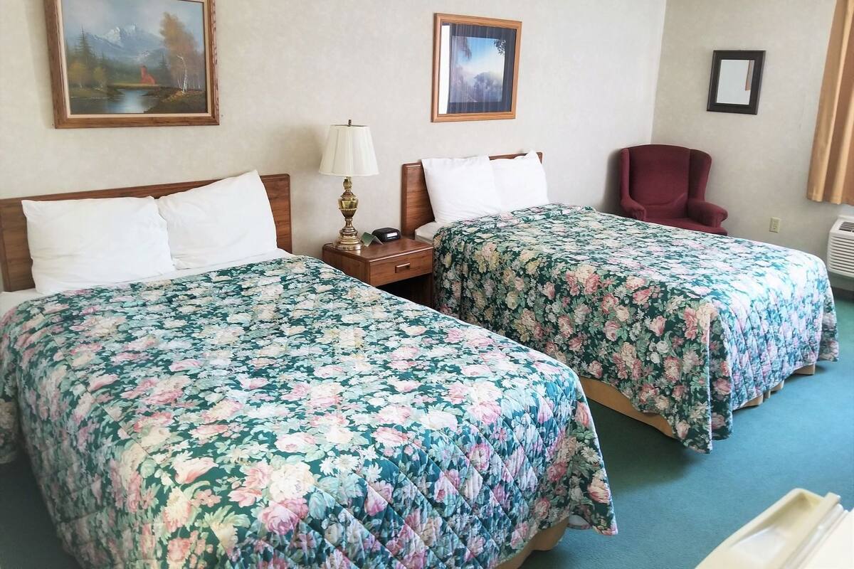 A hotel room with two beds, a nightstand with a lamp, a chair, and two framed pictures on the wall.
