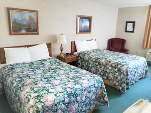 A hotel room with two beds, a nightstand with a lamp, a chair, and two framed pictures on the wall.
