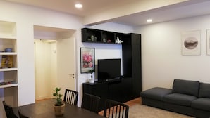 A modern living room with a dark sofa, a dining table with chairs, a TV on a black stand, and a bookshelf with books and decorative items. - Republica83-Campo Pequeno Home (Lisbon)
