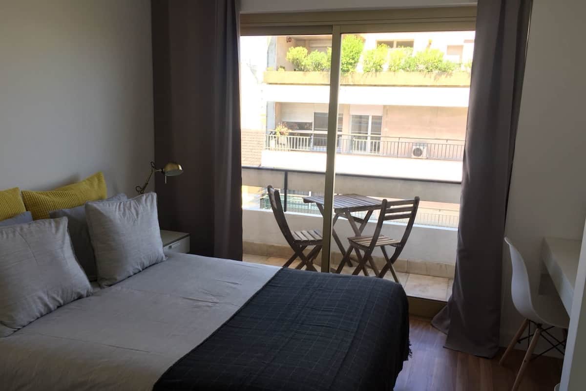 A bedroom with a bed, a nightstand, a desk, and a chair. There is a balcony with a table and chairs.