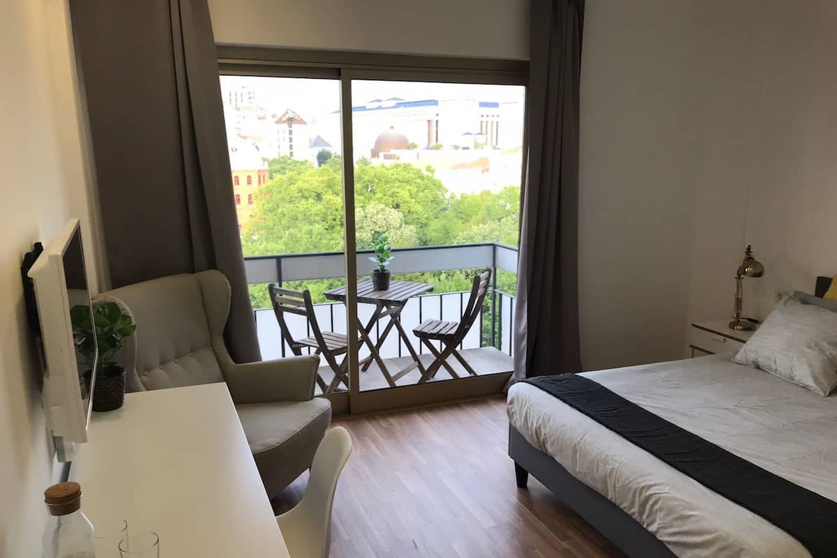 A hotel room with a bed, a desk with a chair, a small table with chairs, a balcony with a view of buildings and trees, and a lamp on the bedside table.